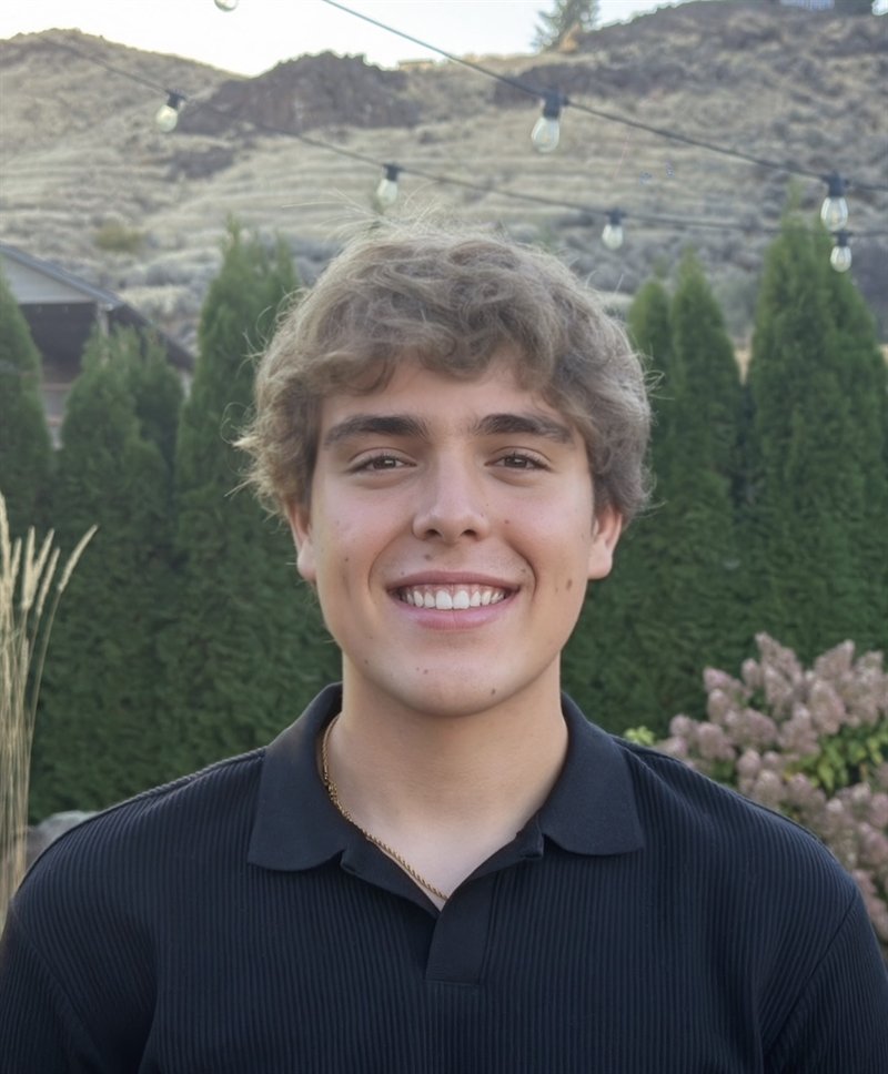 Noah Rogers smiling outdoors in a black polo, with evergreen trees and a hillside behind him