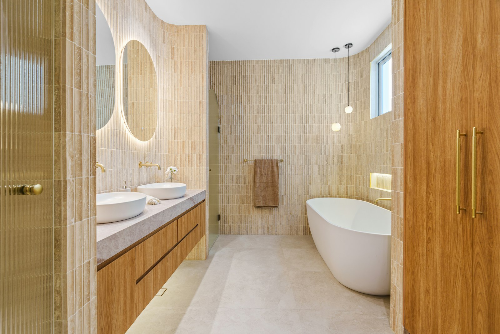 A warm primary bathroom with a freestanding tub, wood cabinetry and round mirrors