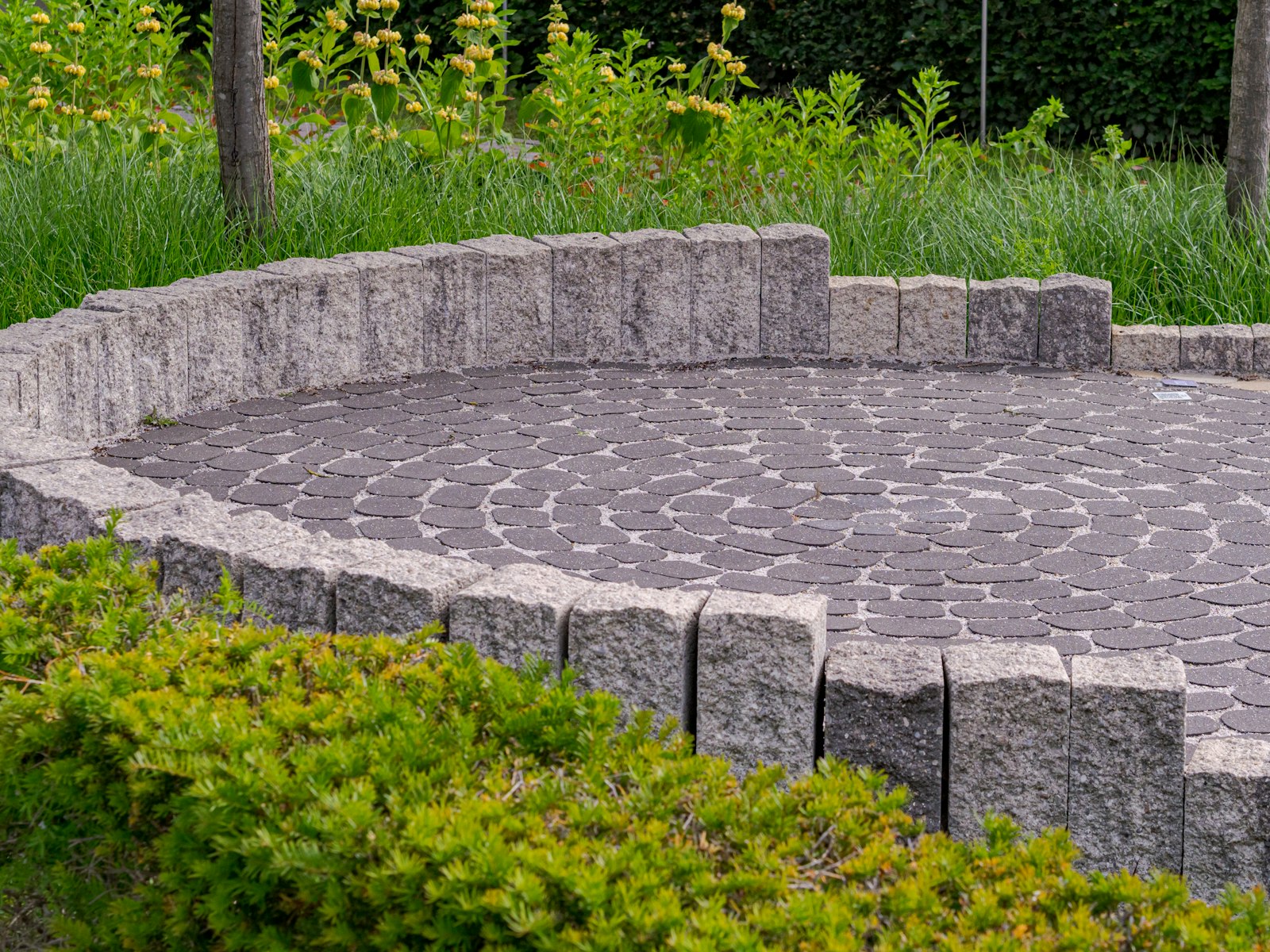 A circular paver patio ringed by a low stone wall, shrubs and grasses